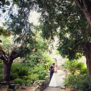 A man and woman dressed in formal wedding clothes embrace and kiss among trees, ferns and ruined walls