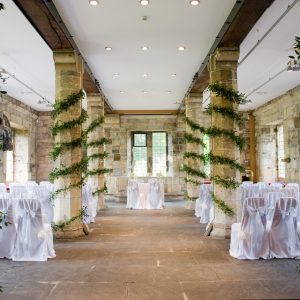 A stone hall with four pillars wrapped in greenery. At the head is a table prepared for a wedding.