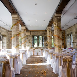 A stone hall with four pillars wrapped in greenery. At the head is a table prepared for a wedding.