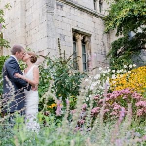 A man and woman in formal wedding attire embrace and kiss in front of a stone building. Spring flowers are all around.