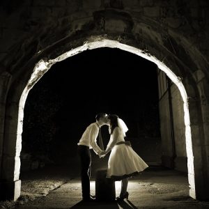 Bride and groom kiss under stone arch at night